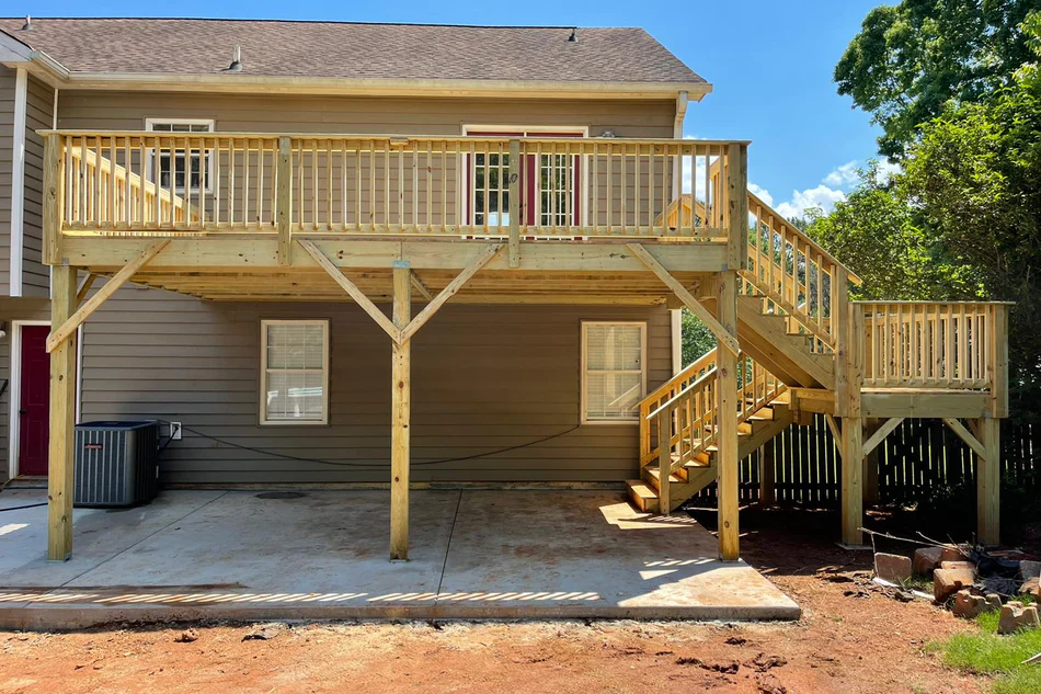 Completed elevated wooden deck with spindle railings, support bracing, and stairs leading to a concrete patio at the rear of a home.